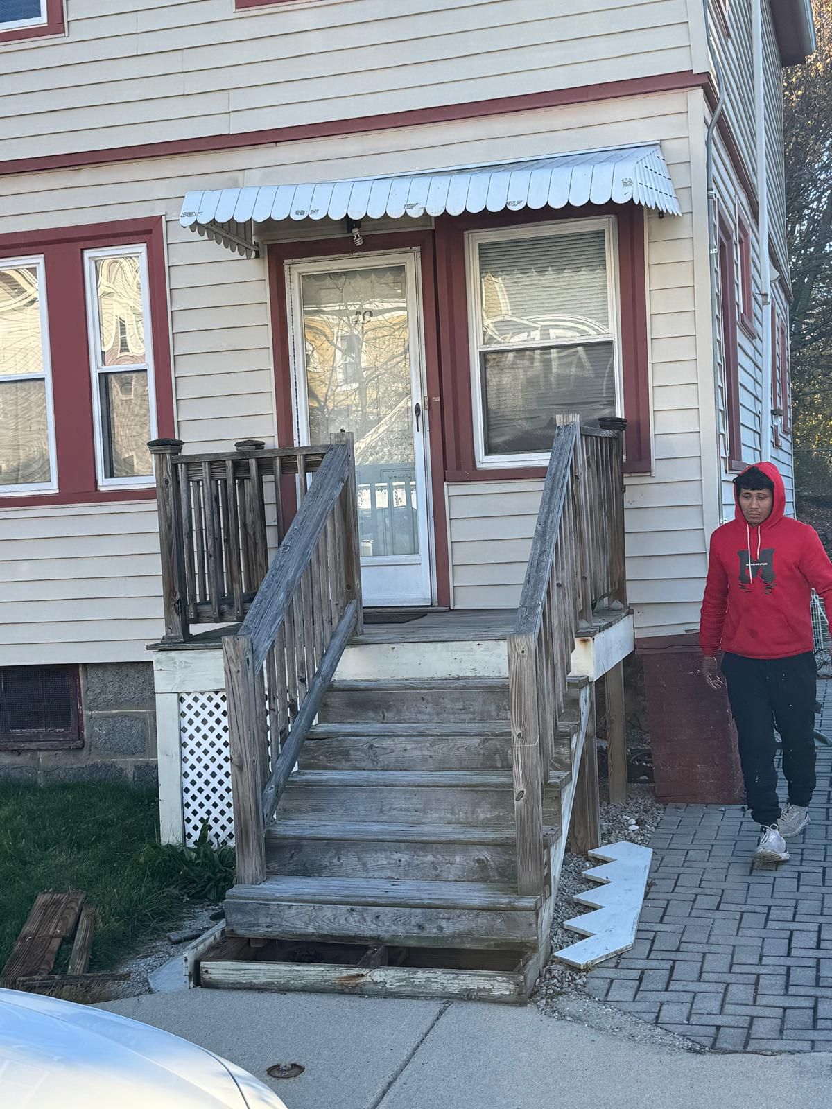 A person in a red hoodie walks past wooden steps leading to the front door of a light-colored, multi-story house.