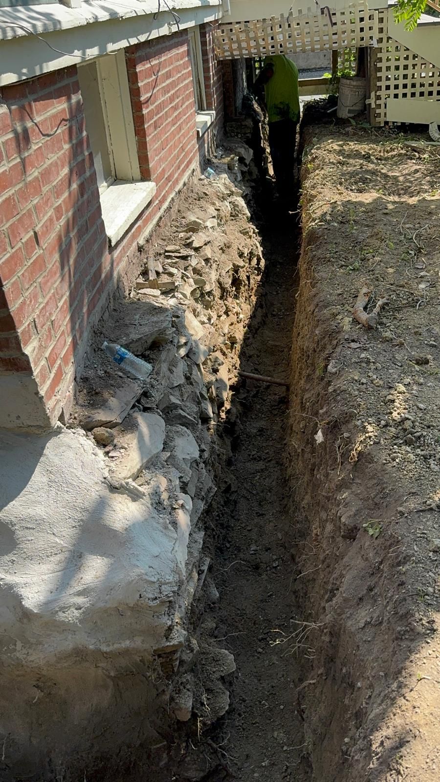 A narrow trench dug along the brick foundation of a house, with a worker visible in the background.
