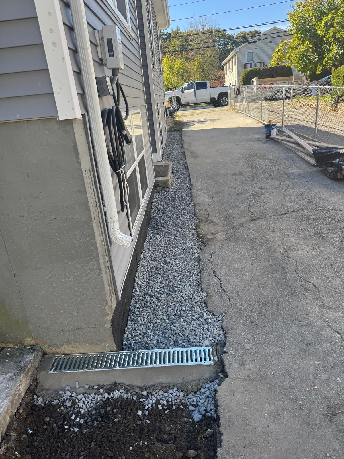 A trench drain and gravel bed installed along the foundation of a gray house exterior next to a cracked asphalt driveway.