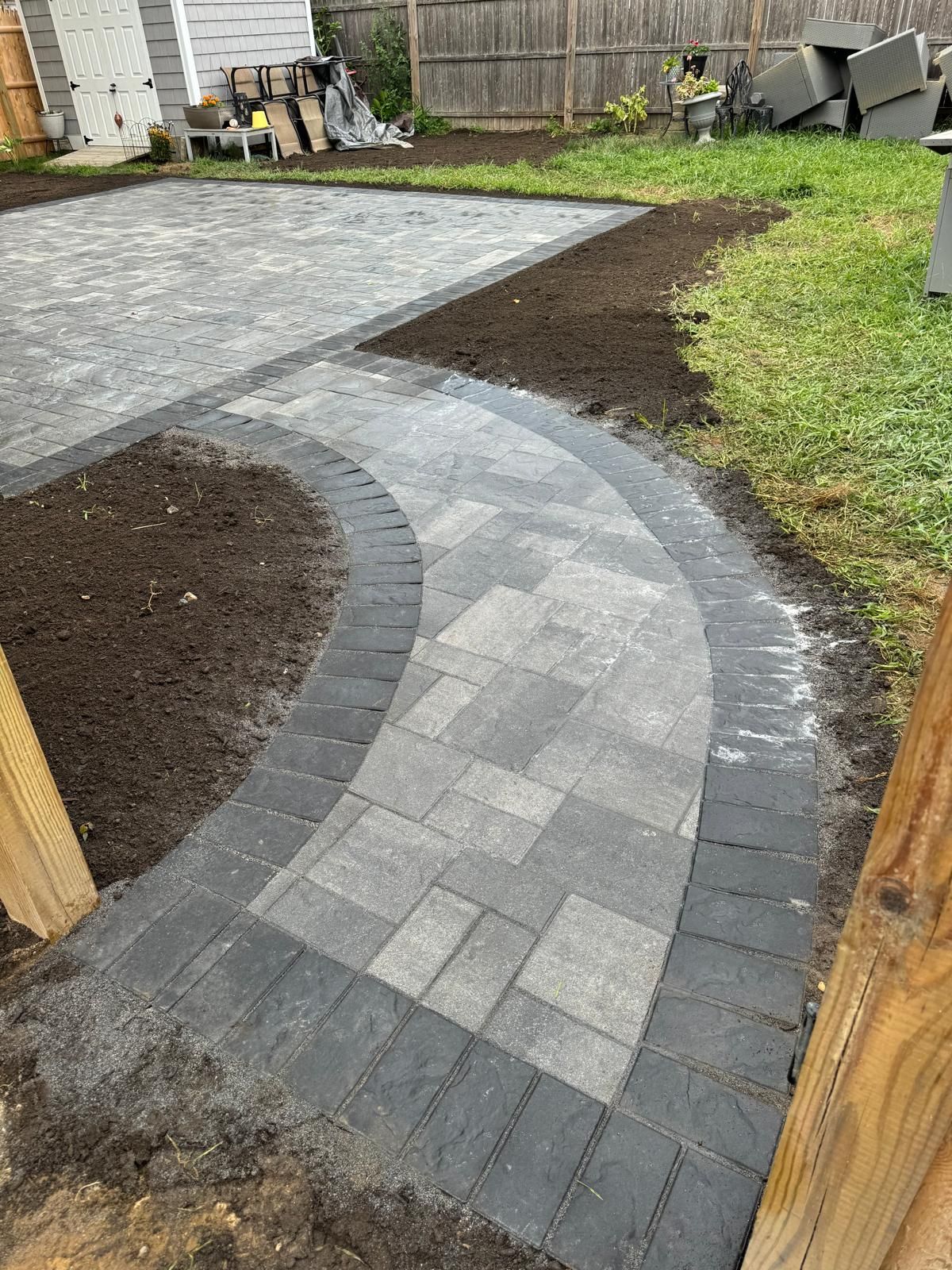 Curved gray paver walkway with a dark border leading to a larger patio area in a backyard under construction.
