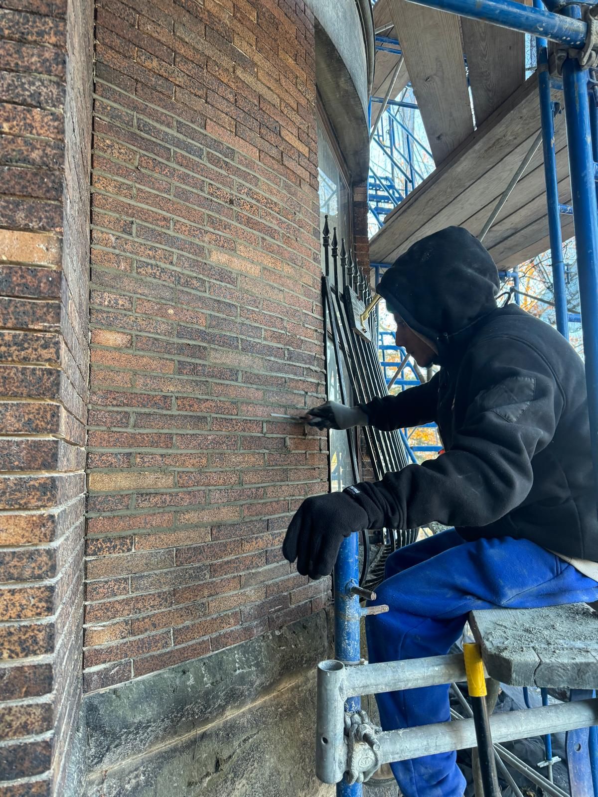 A worker in a black hoodie and blue pants applies mortar to a dark brick wall while perched on metal scaffolding.