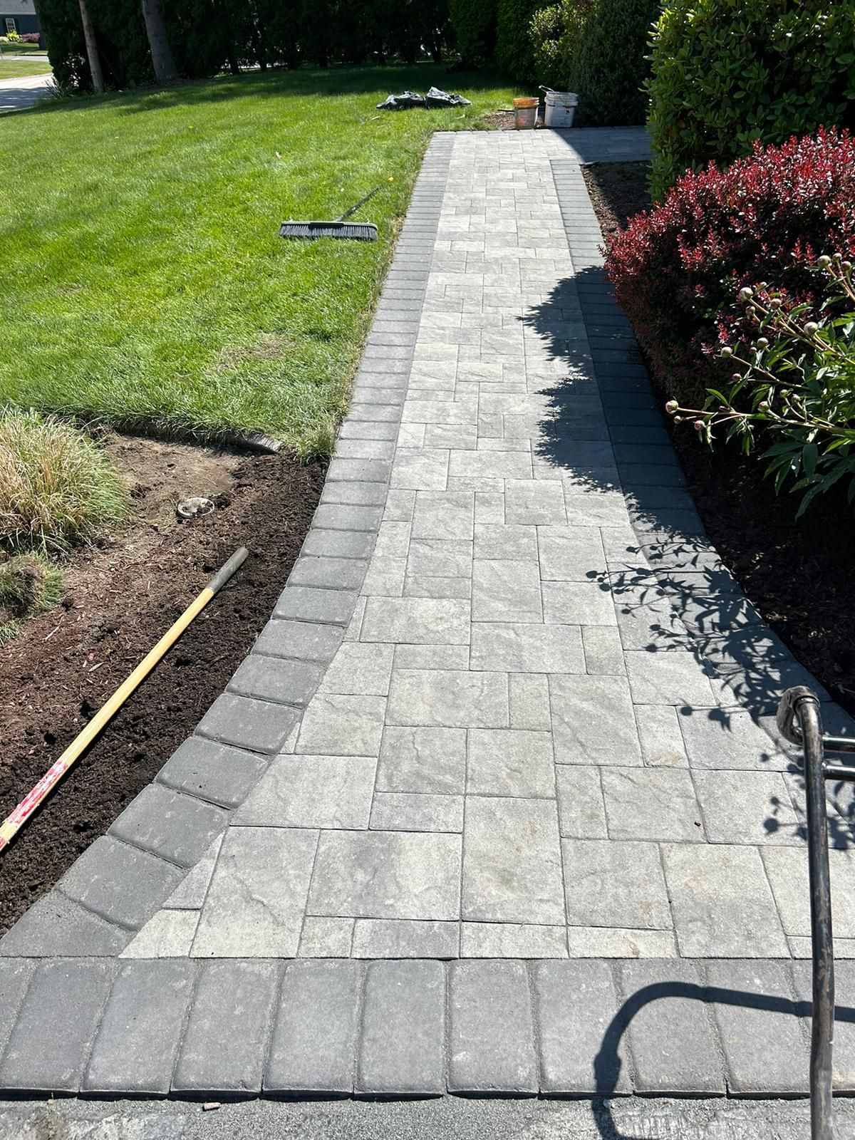 A straight, rectangular stone walkway with a dark border runs through a green lawn and alongside a garden bed.