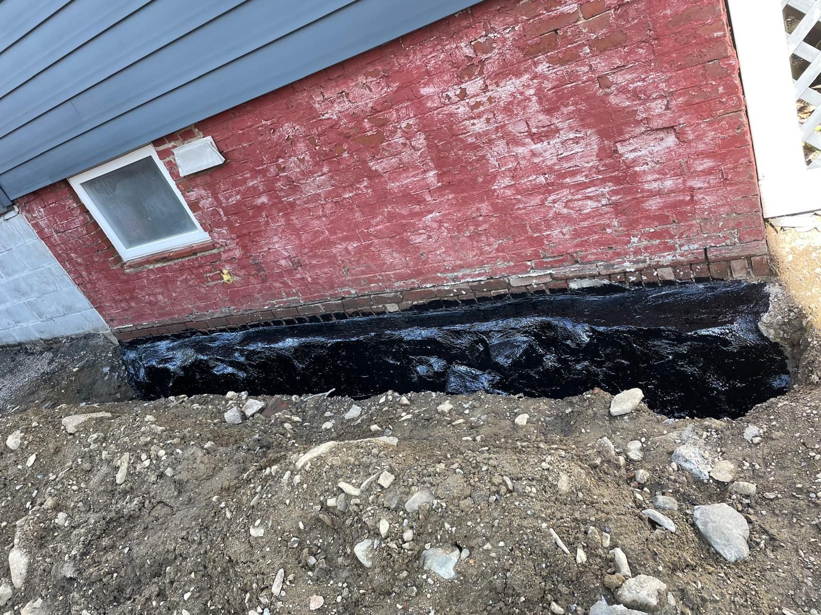 Exterior brick foundation wall coated with black waterproof sealant above a dirt trench.