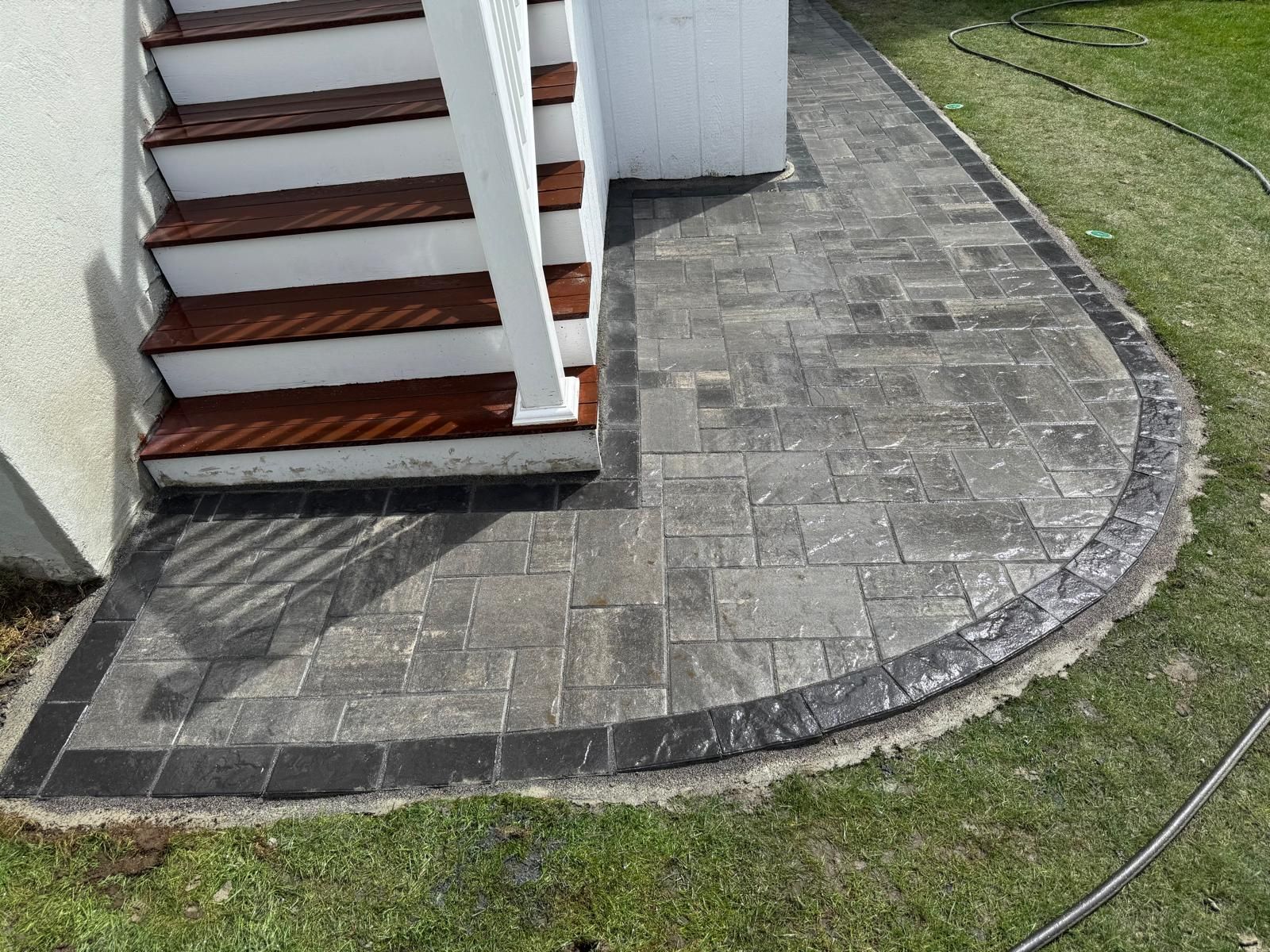 A curved paver patio with a dark border sits at the base of wooden outdoor stairs against a white house wall.
