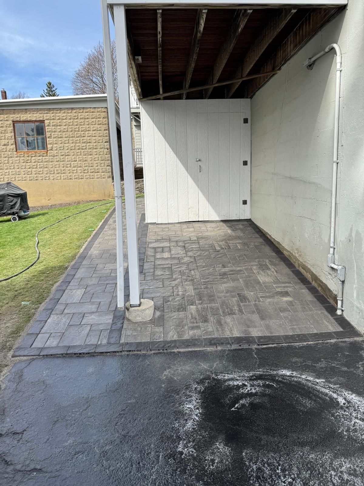 New gray paver patio under a deck, bordered by dark stone, adjacent to a white wall and a black asphalt driveway.
