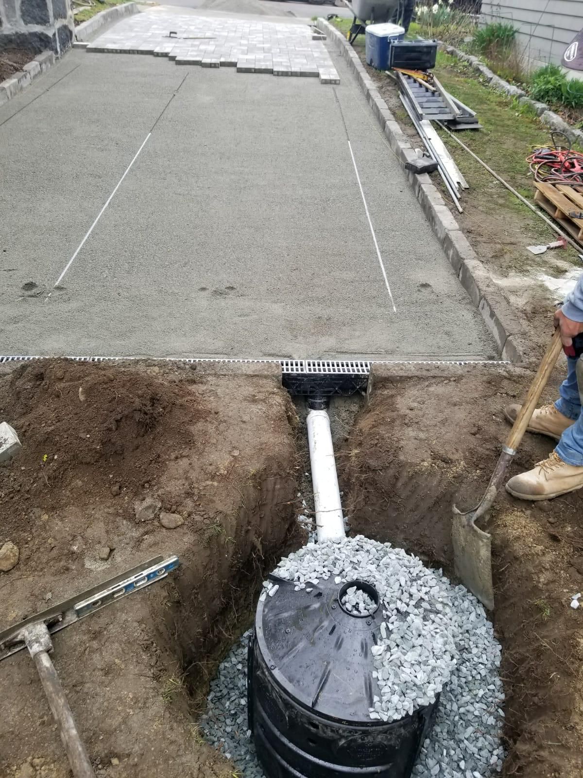 A worker installing a drainage system for a paved driveway, featuring a catch basin connected to a gravel-filled dry well.