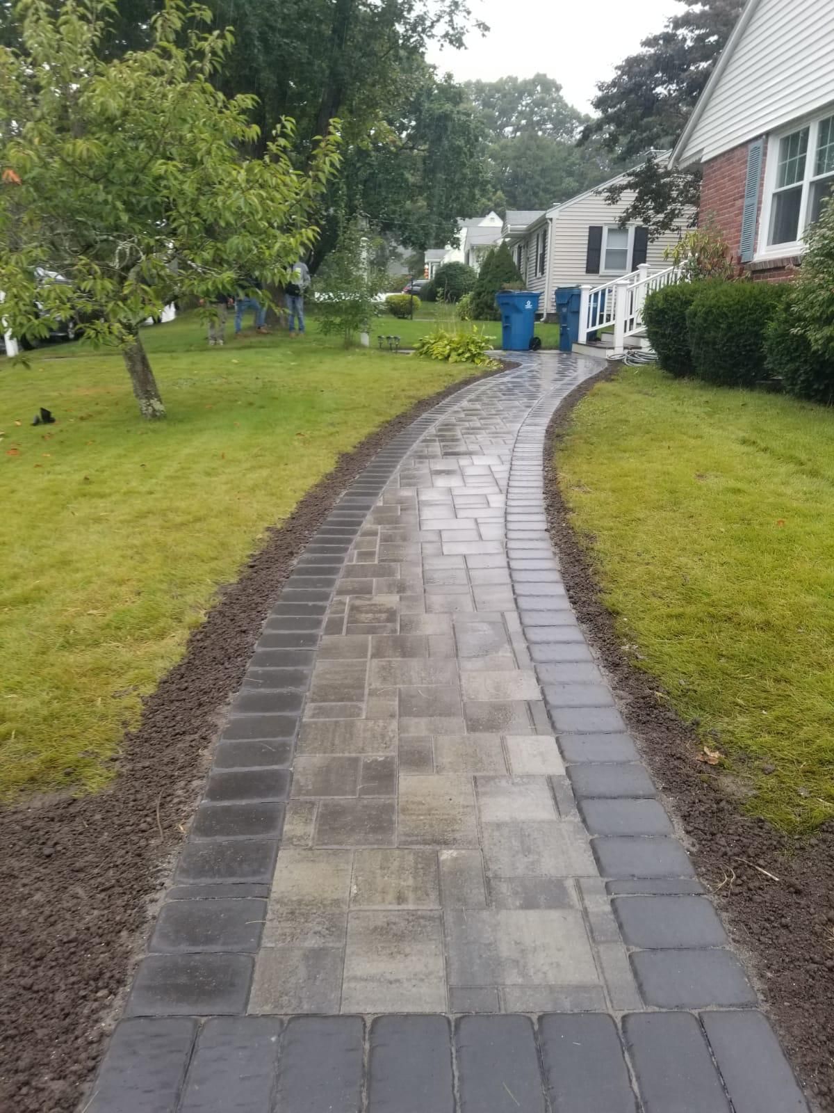 A newly installed paver walkway with a dark border and light center, winding through a suburban front yard.