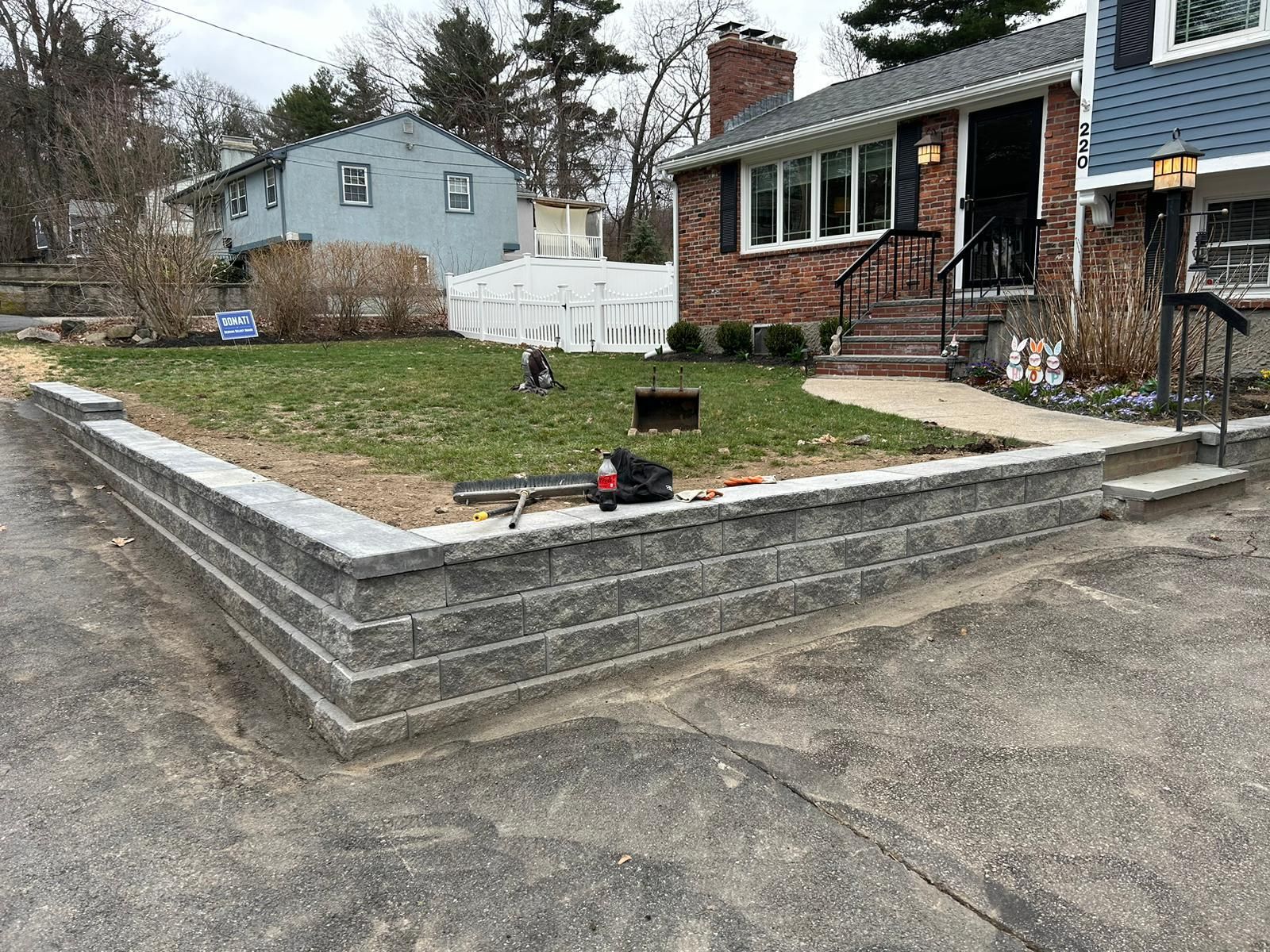 A newly constructed gray concrete block retaining wall borders a front lawn in front of a brick house.