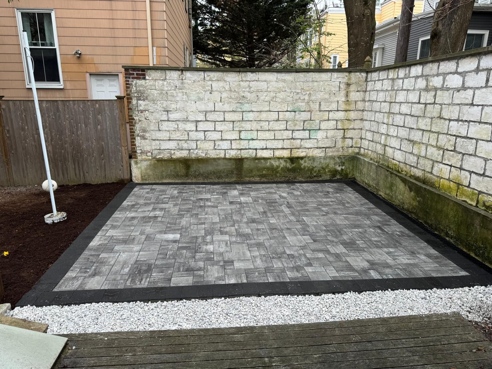 A gray stone patio with a dark border, bordered by white gravel, next to a white brick wall in a backyard setting.