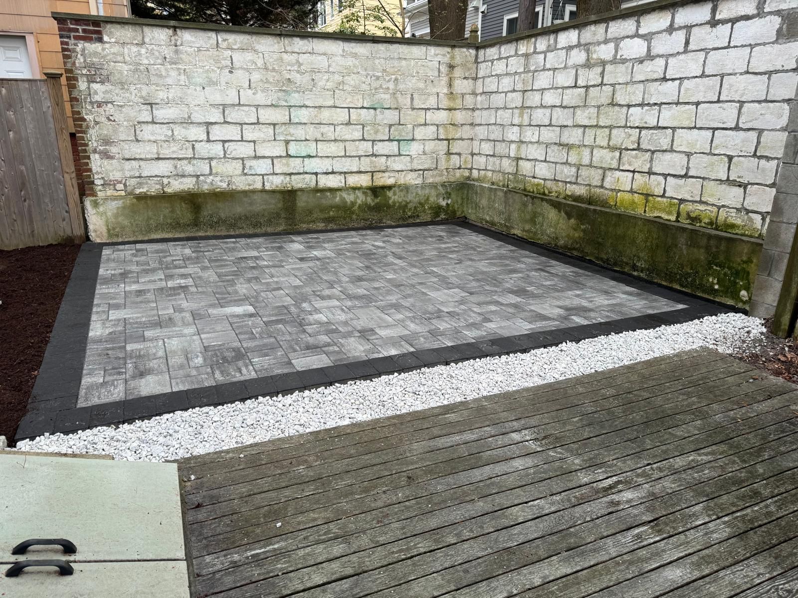 A gray stone patio with a dark border, bordered by white gravel and a wooden deck, set against a weathered brick wall.
