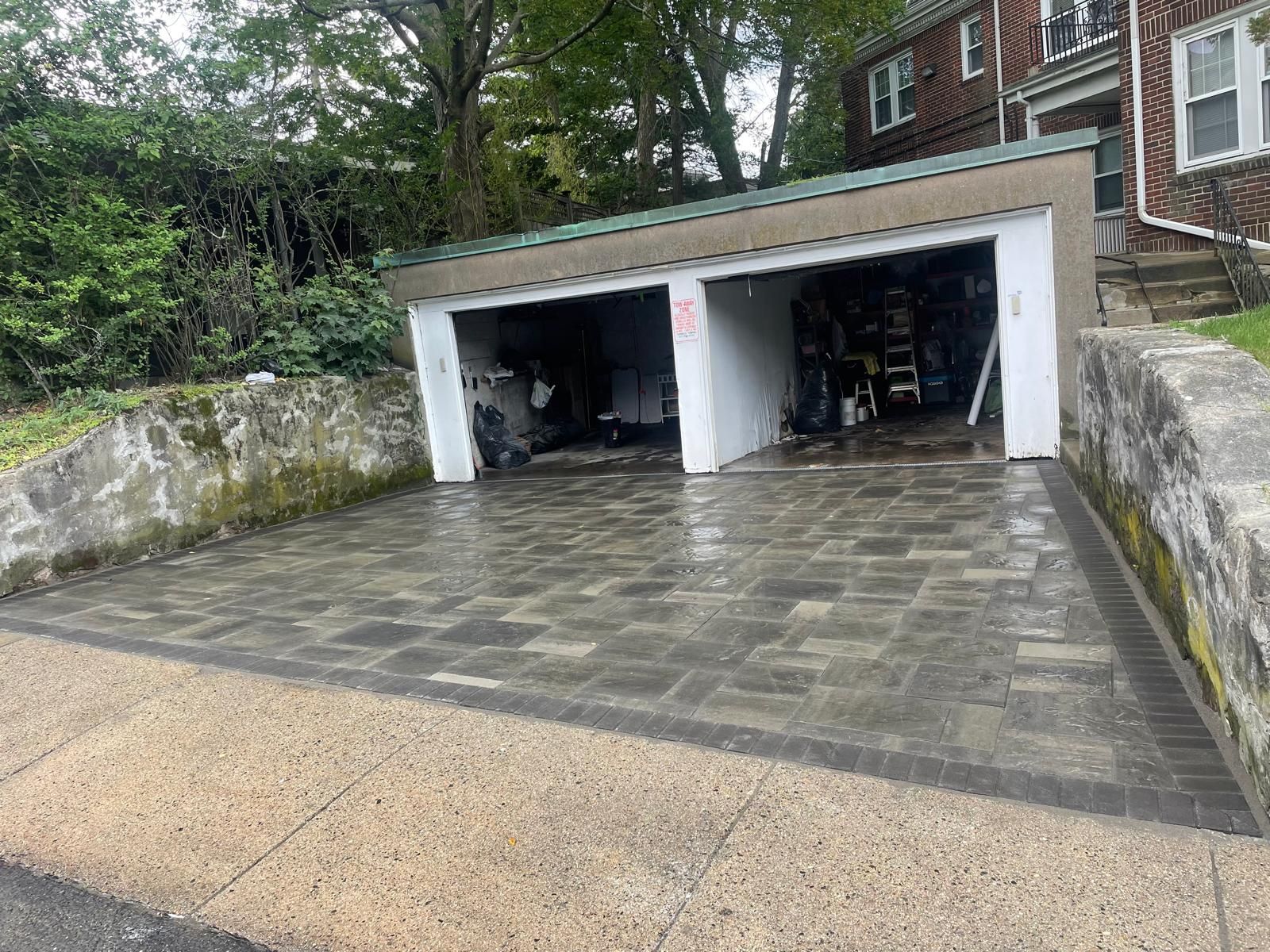 A two-car garage with a newly paved paver driveway, framed by low stone retaining walls and green trees.