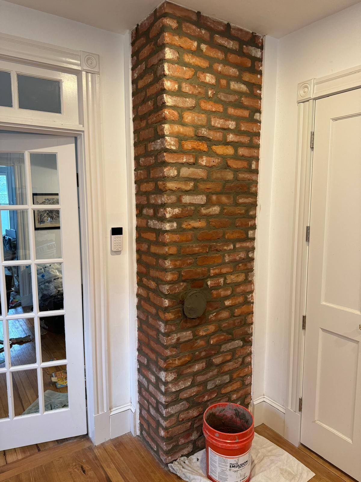 An exposed brick chimney column stands in a hallway between a white paneled door and a glass-paned door, with a paint bucket.