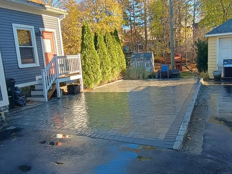 A gray house with a wooden staircase leading to a new, rectangular stone paver patio in a residential backyard.