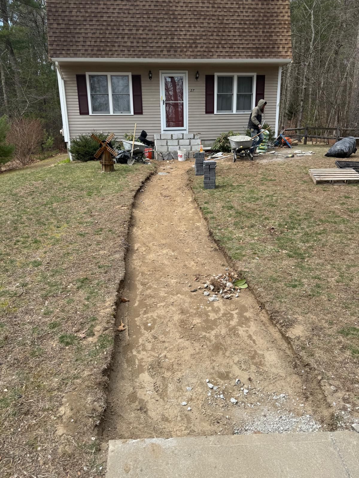 A person works on a dirt walkway path leading to the front door of a suburban house on a sunny day.