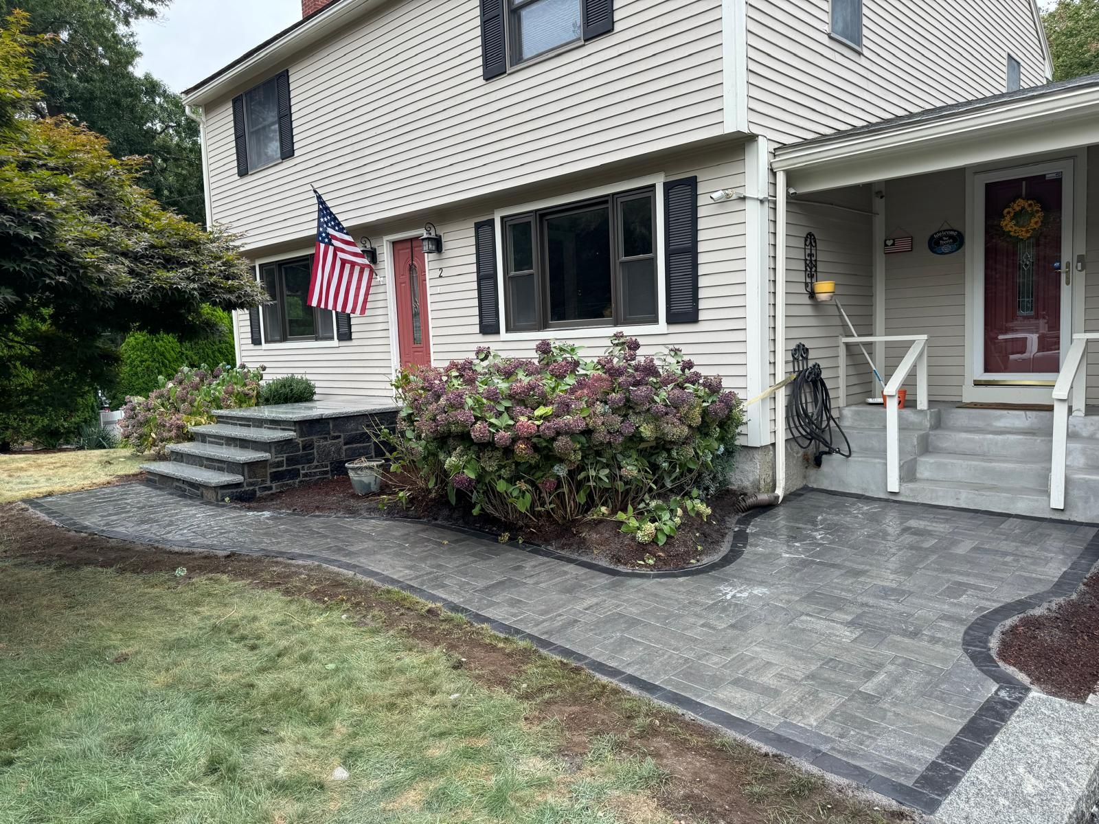 A front yard landscape with a newly installed gray stone patio and walkway leading to a house with beige siding.