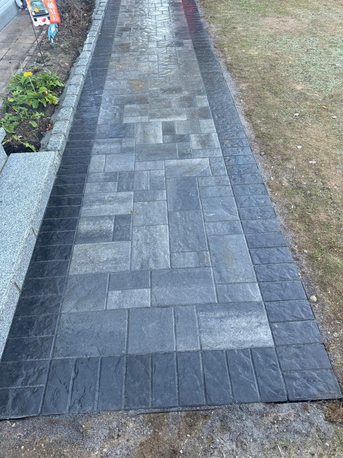A straight outdoor walkway made of gray rectangular pavers with a dark charcoal border, flanked by grass and a planter.