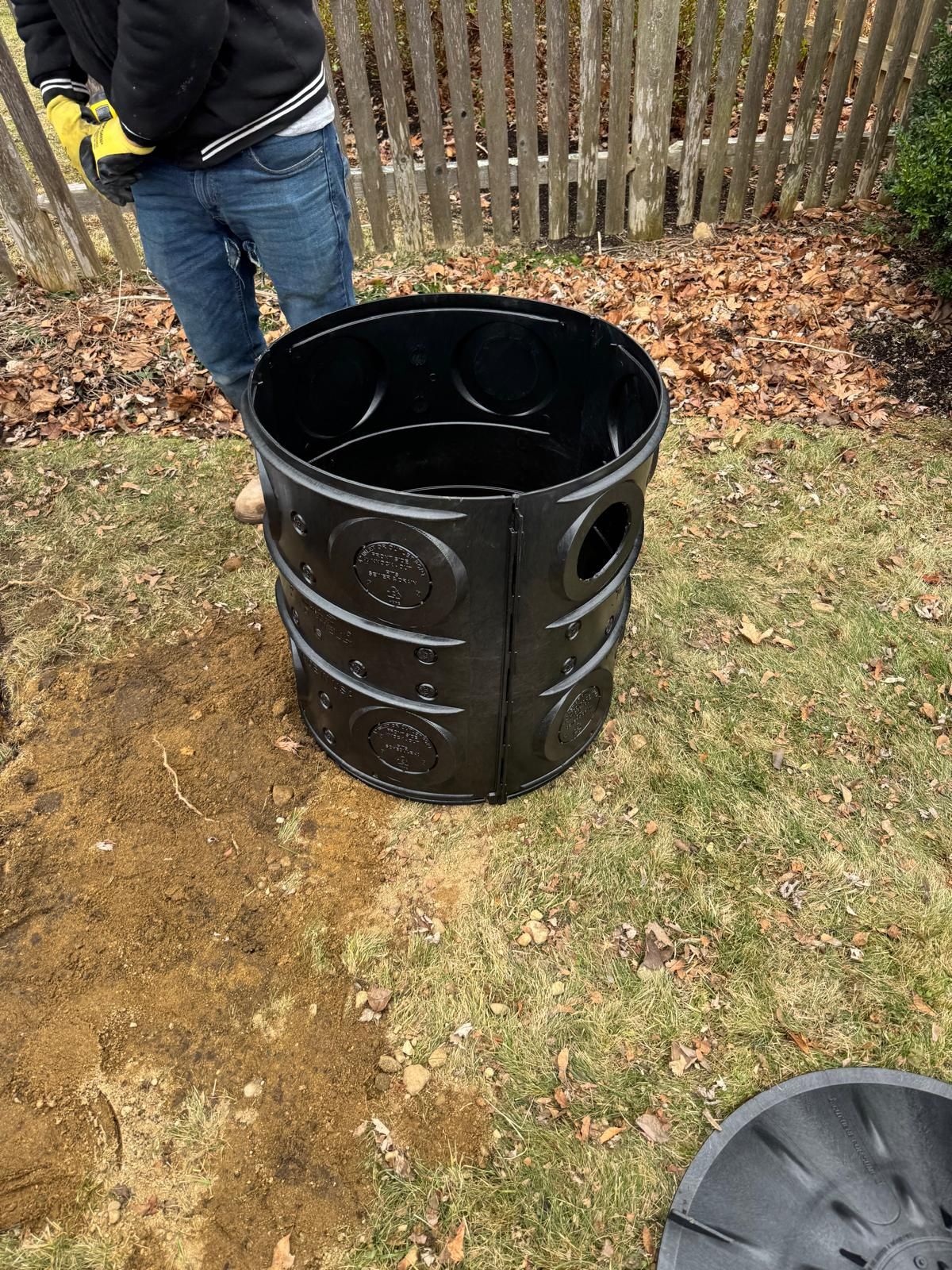 A black plastic drainage basin stands on grass in a yard next to an open hole, with a person wearing work gloves nearby.