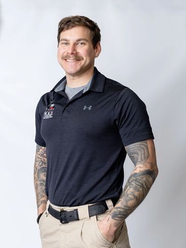 Smiling man in a navy polo and khaki pants with tattooed arms against a light gray background