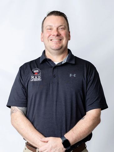 Smiling man in navy polo shirt standing with hands clasped against a light gray background