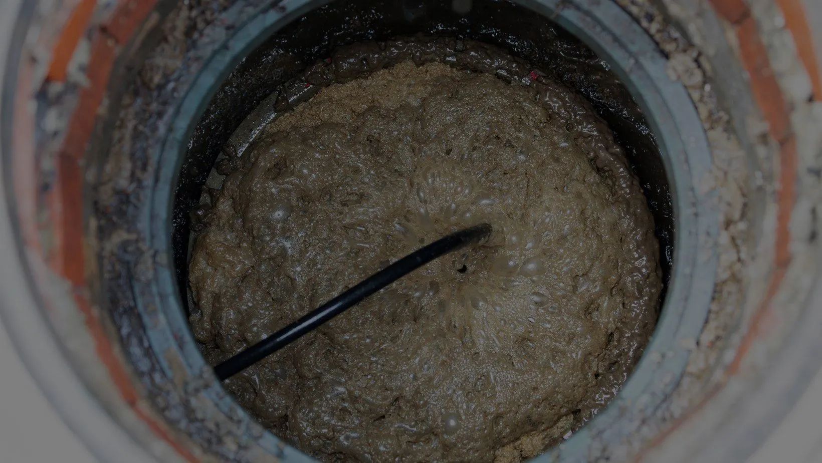 Top view into a narrow pipe with a black rod crossing a circular opening filled with beige sediment.