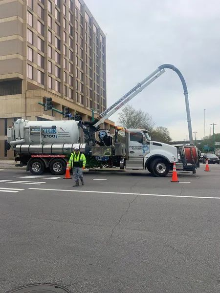 Utility truck with boom hose parked beside a building, with workers and orange cones on the street.