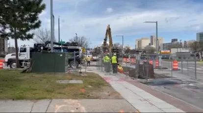 Road construction scene with workers, excavator, orange barriers, and sidewalk along a wet street