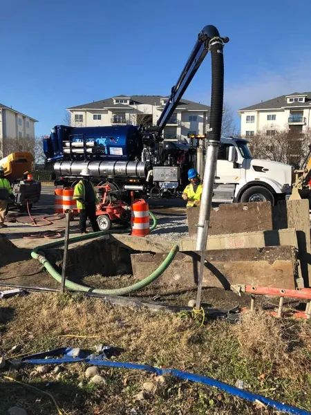 Construction crew operating a white vacuum truck at a roadside work site with hoses and orange cones