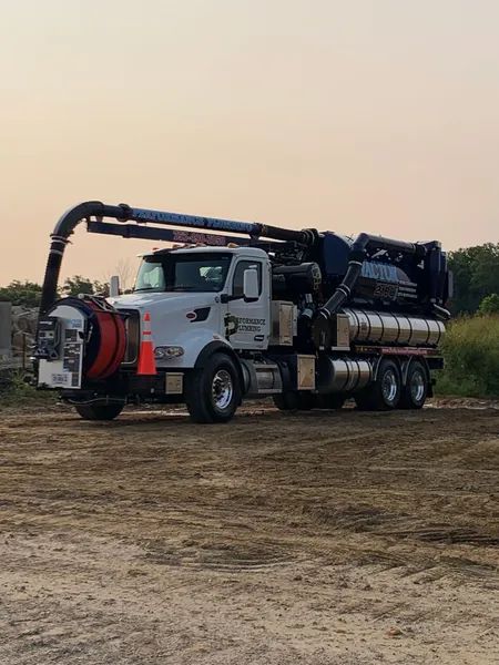 White vacuum truck with a large hose parked on a dirt field at dusk.