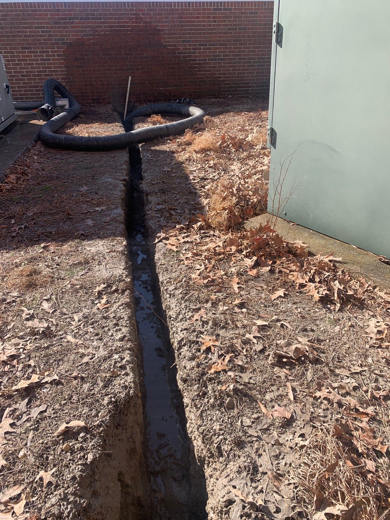 Narrow trench cut through dirt beside a brick wall, with a black hose lying across the top.