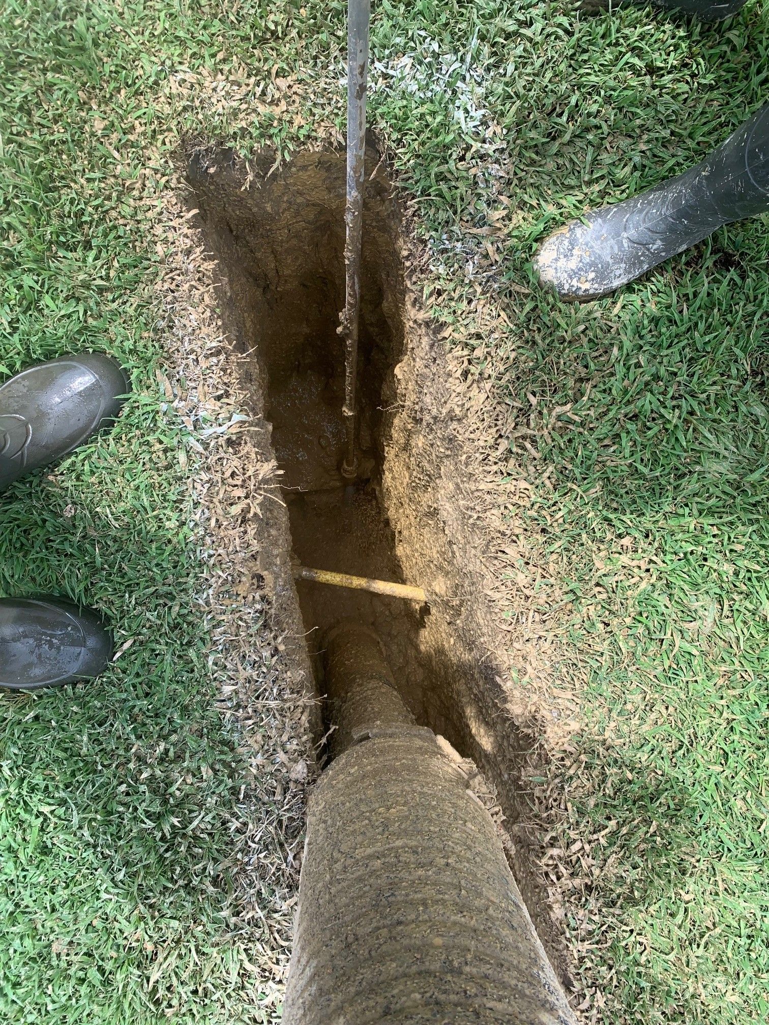 Two people stand beside a narrow trench exposing a buried utility pipe in grass.