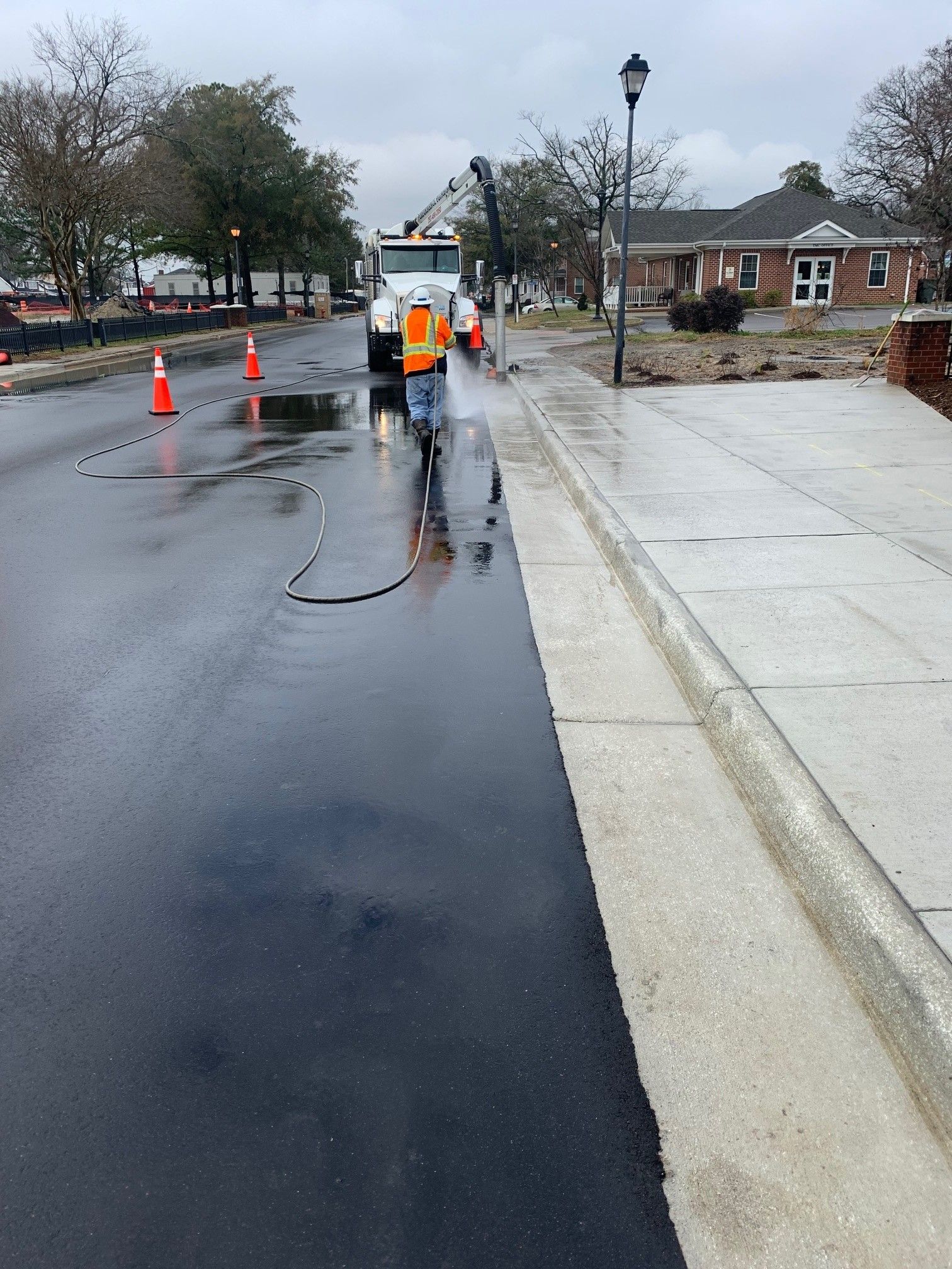 Worker in orange vest with hose on wet street near sidewalk cones and curb ramp