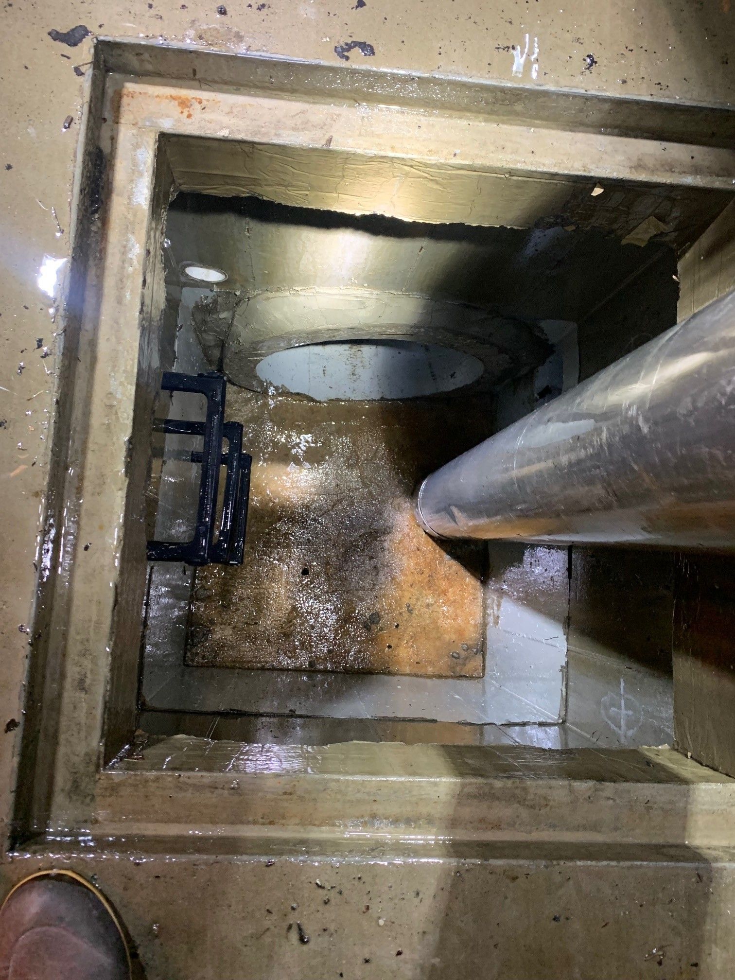 Looking down a square concrete shaft with a pipe, circular opening, and metal ladder rungs.