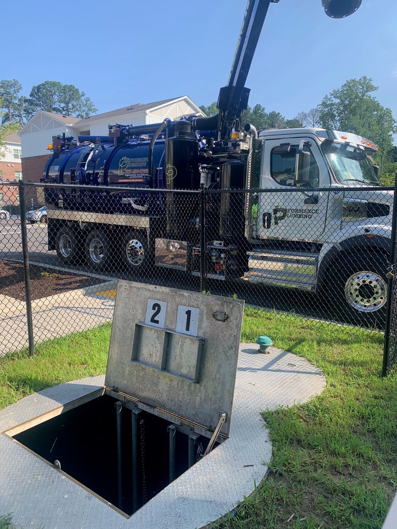 Truck with crane over open utility vault in a grassy area, beside a chain-link fence.