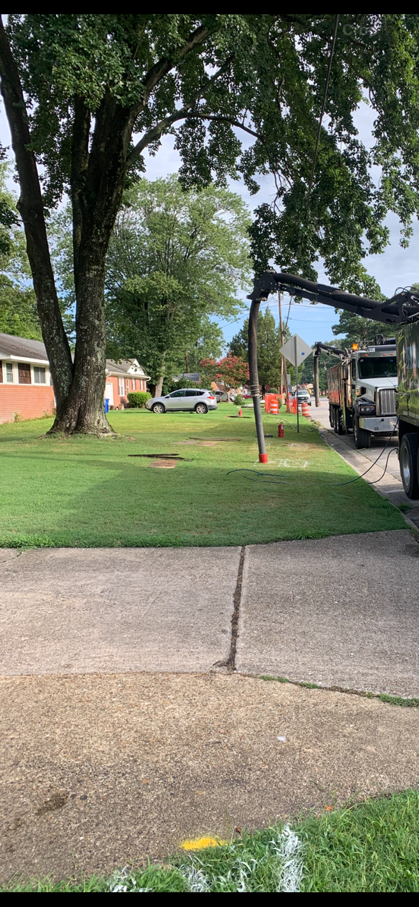 Sidewalk under large trees in a grassy neighborhood, with a red-post sign and parked vehicles nearby.
