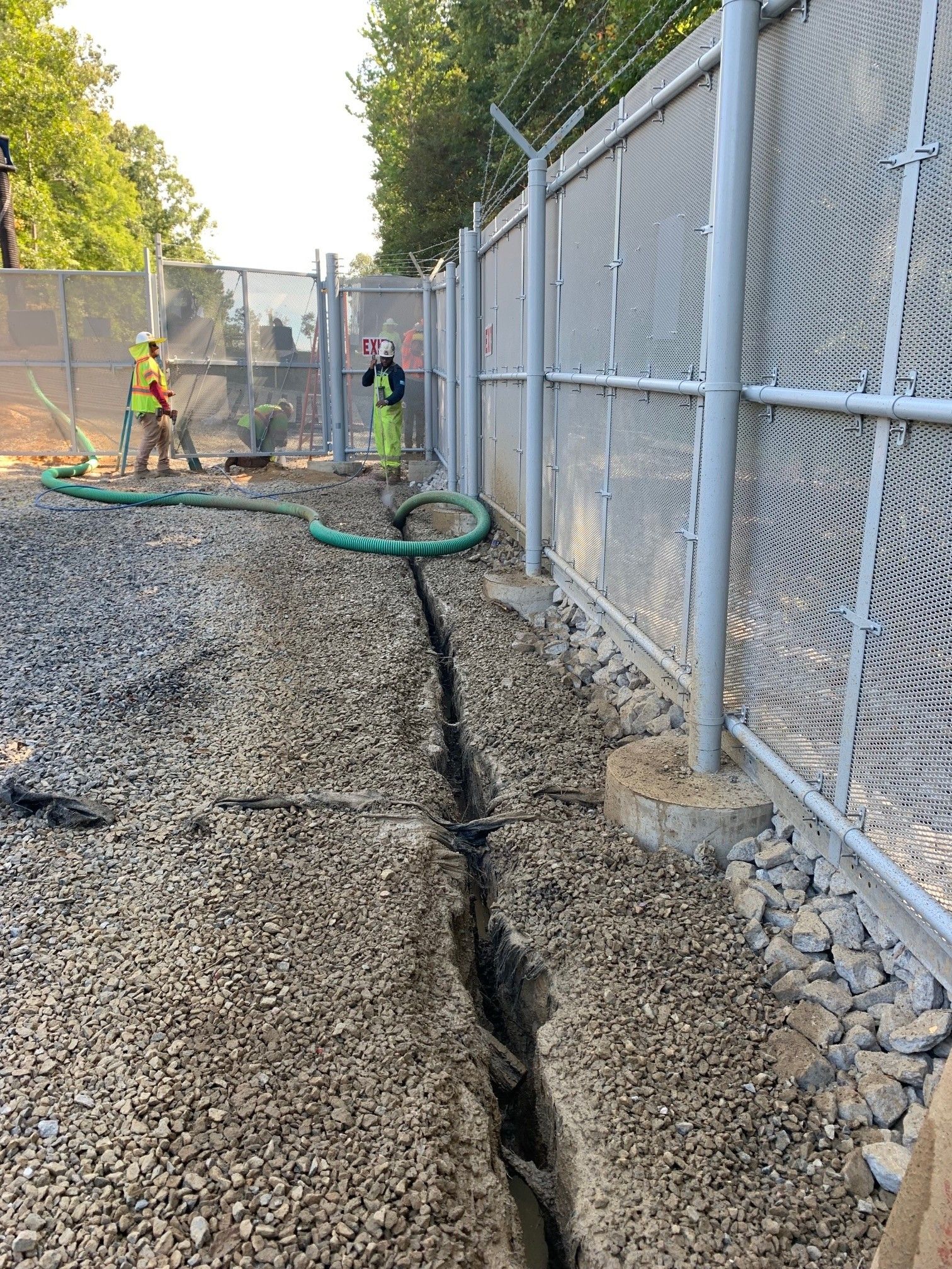 Workers repairing a gravel trench beside a chain-link fence, with a hose laid across the ground.