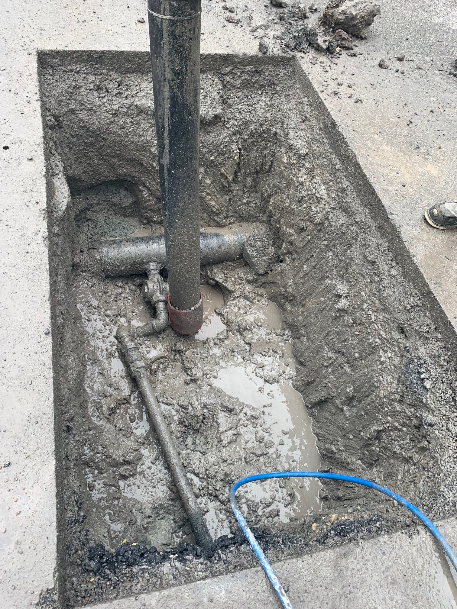 A broken concrete trench with exposed pipes and a vertical metal pole, with a blue cable on the ground.