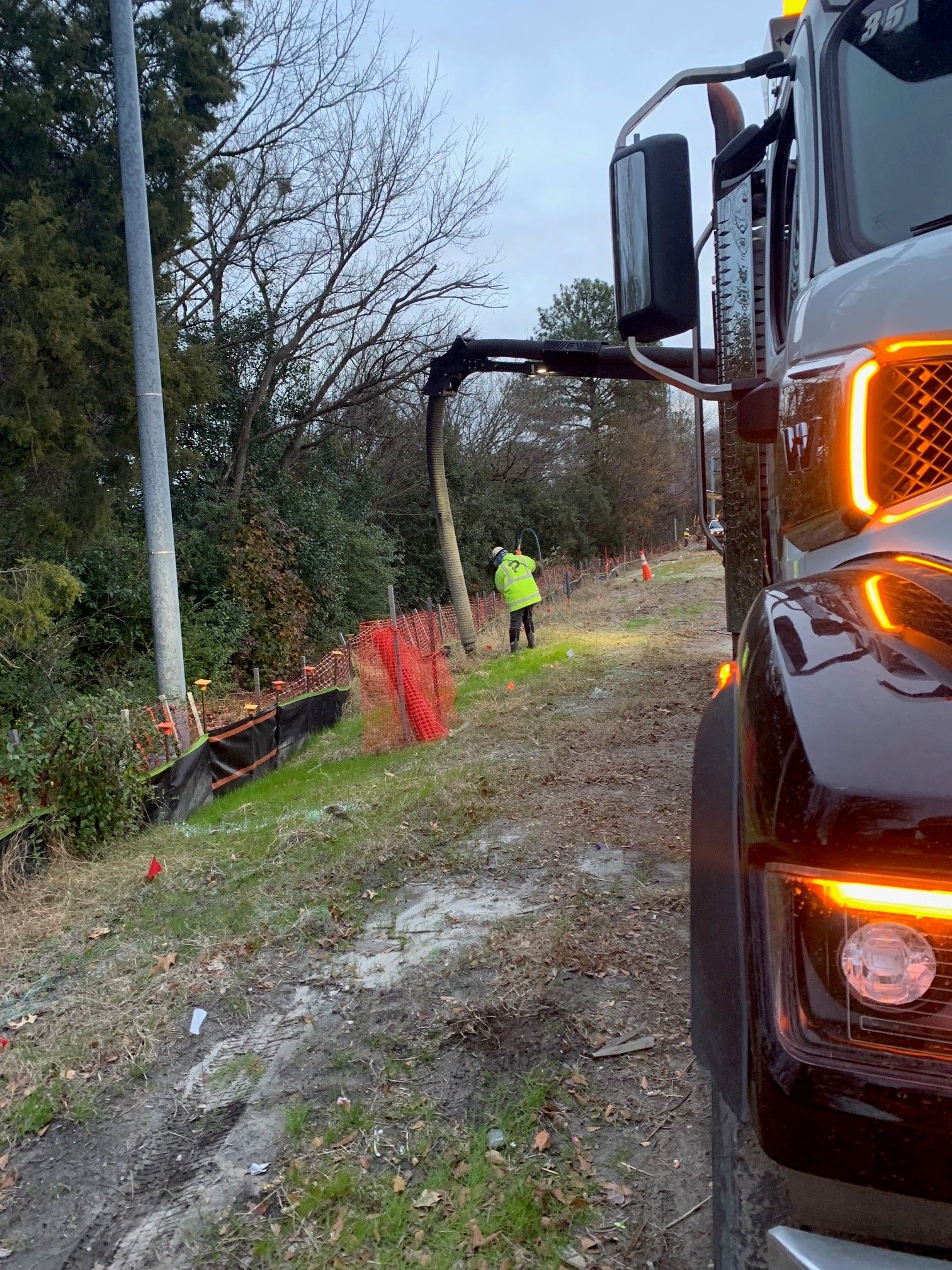 Tow truck on muddy roadside beside a worker in a neon jacket, with orange safety fencing and trees nearby