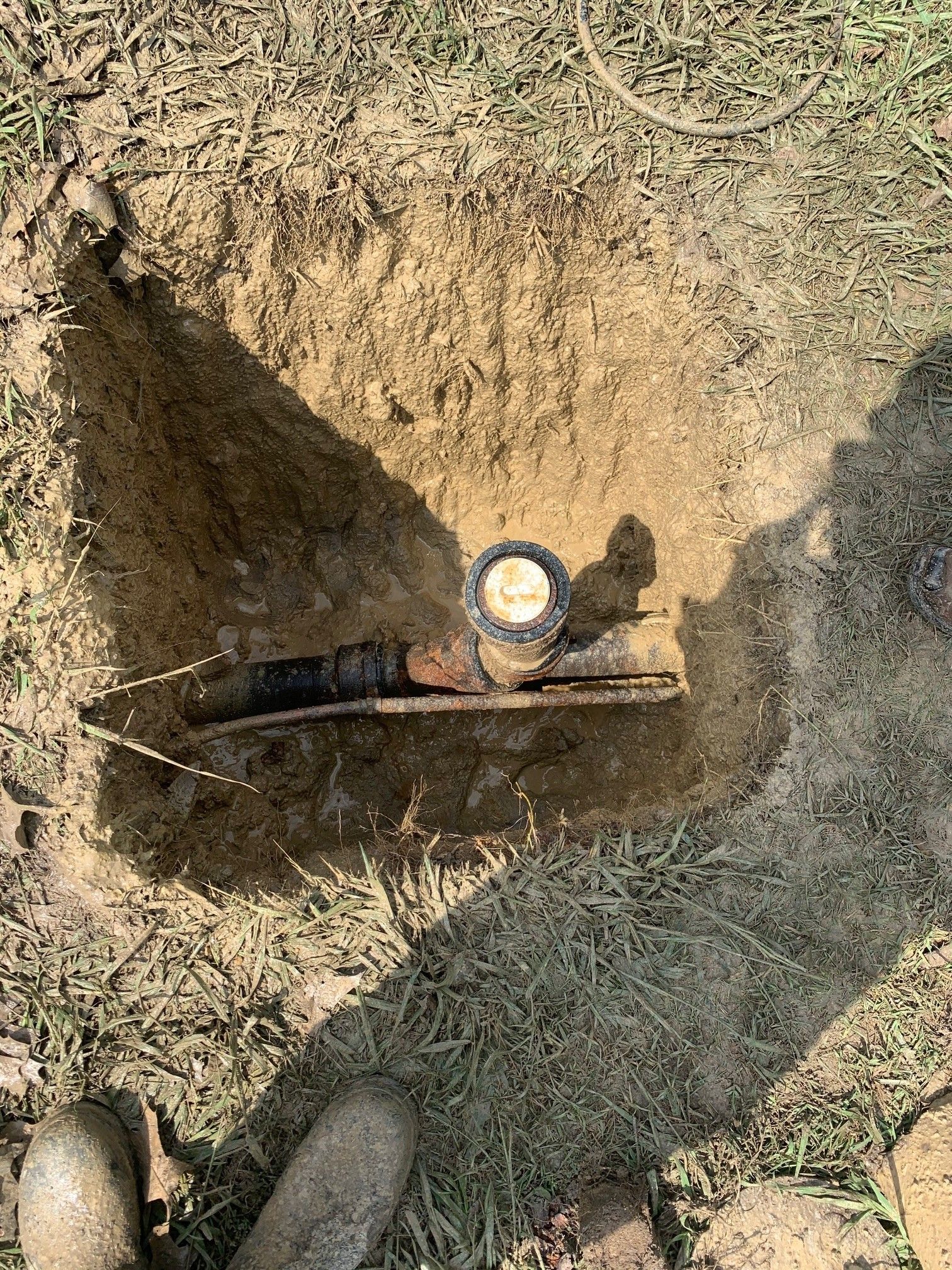 Excavated dirt hole with a pressure gauge and valve attached to a pipe, boots visible at bottom left.