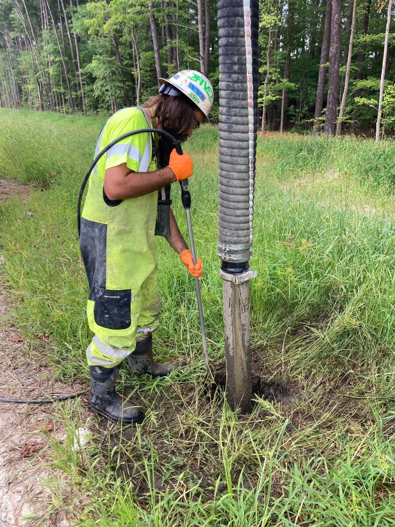 Worker in neon safety gear trimming grass around a wooden utility pole in a wooded field