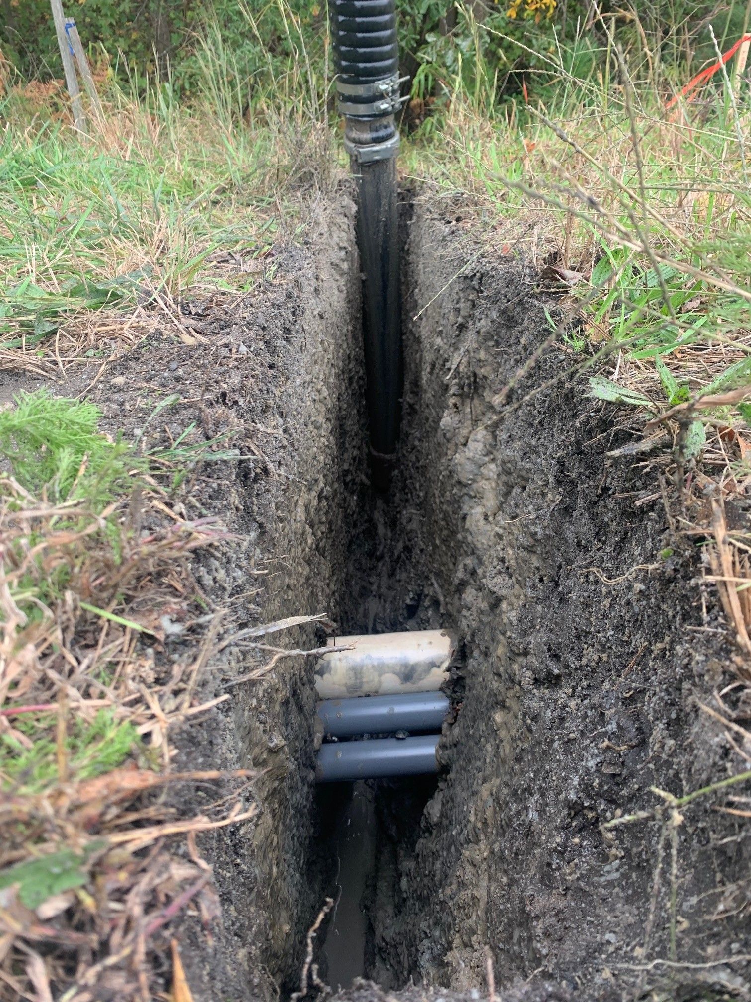 A narrow trench in soil with exposed utility pipes and a black conduit running vertically through the center.