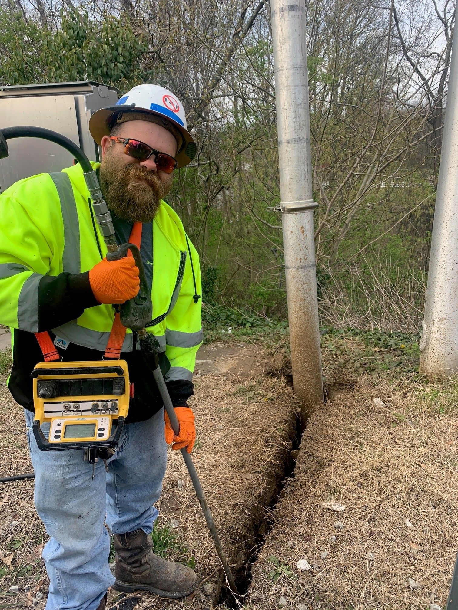 Worker in hard hat and safety vest using a utility locator beside a dug trench outdoors