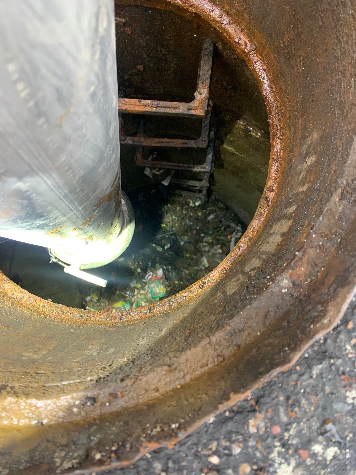 Open manhole with rusty rim and a metal pipe inside, showing standing water and debris.