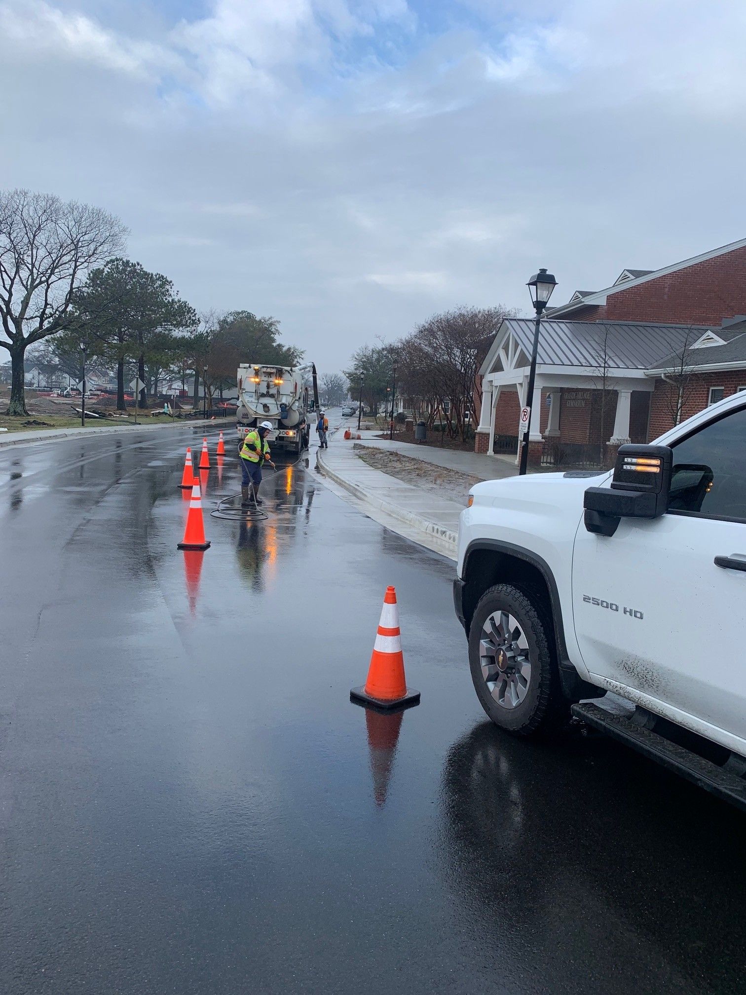 Workers and orange cones line a wet street beside a white utility truck on a rainy day.