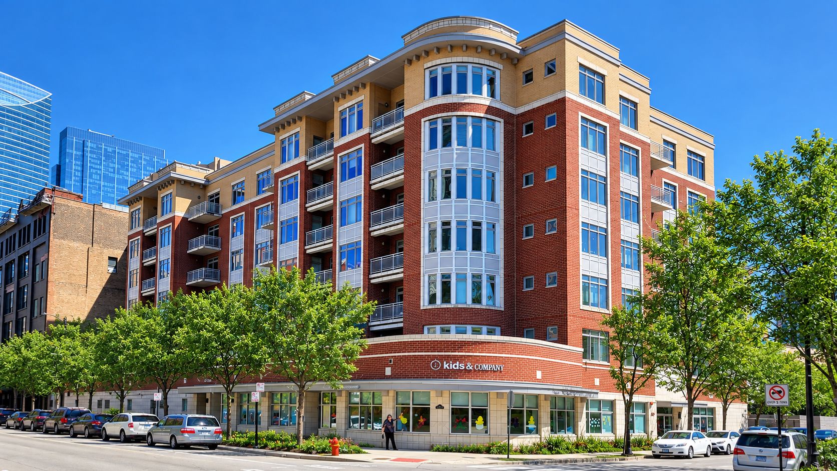 Multi-story brick apartment building with rounded corner, ground-floor shops, and parked cars on a sunny street.