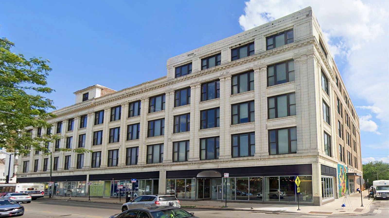 Corner street view of a beige multi-story building with storefronts and parked cars on a sunny day