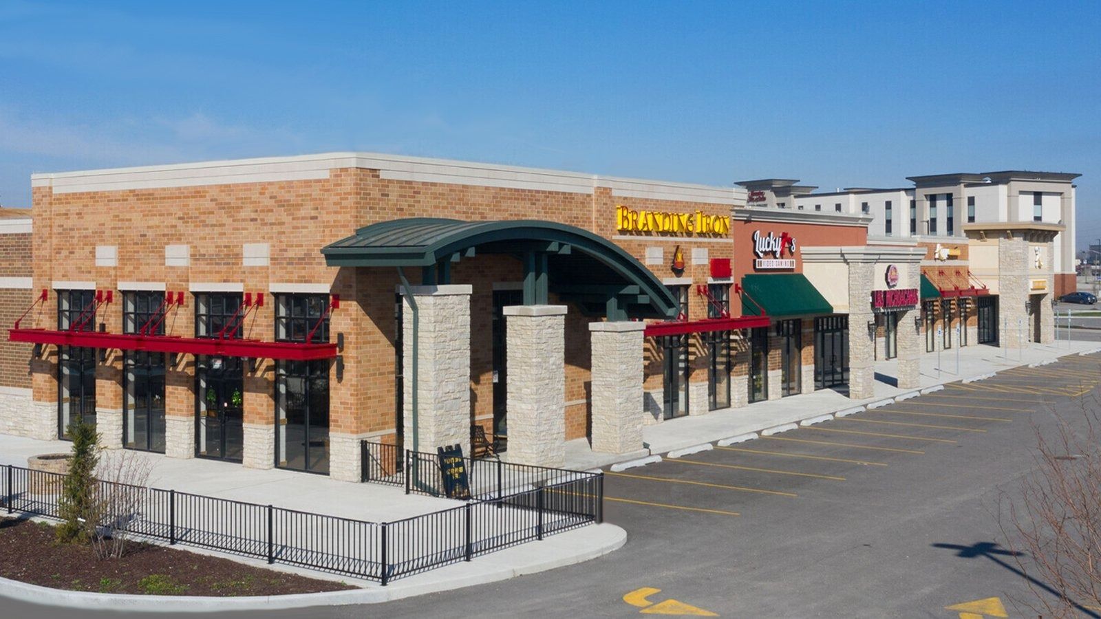 Brick storefront shopping plaza with red awnings and a green entrance canopy on a sunny street corner