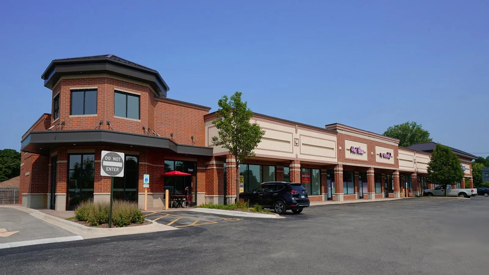 Strip mall storefronts with beige facades and a corner brick building under a clear blue sky