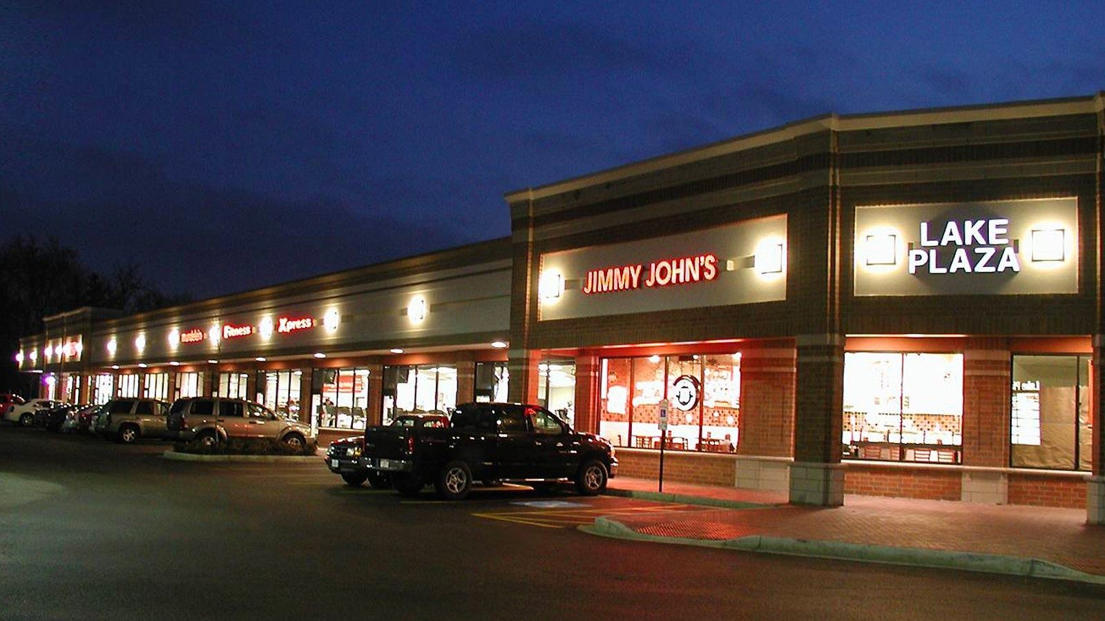 Nighttime strip mall with lit storefronts, including a DAVEY JONES and LAKE PLAZA sign, and parked cars.