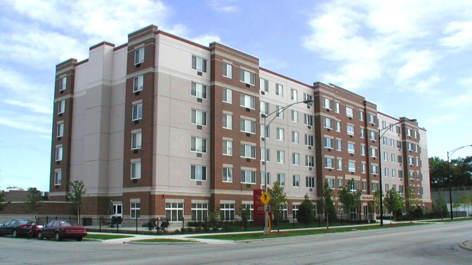 Six-story brick and beige apartment building on a city street corner under a cloudy sky.