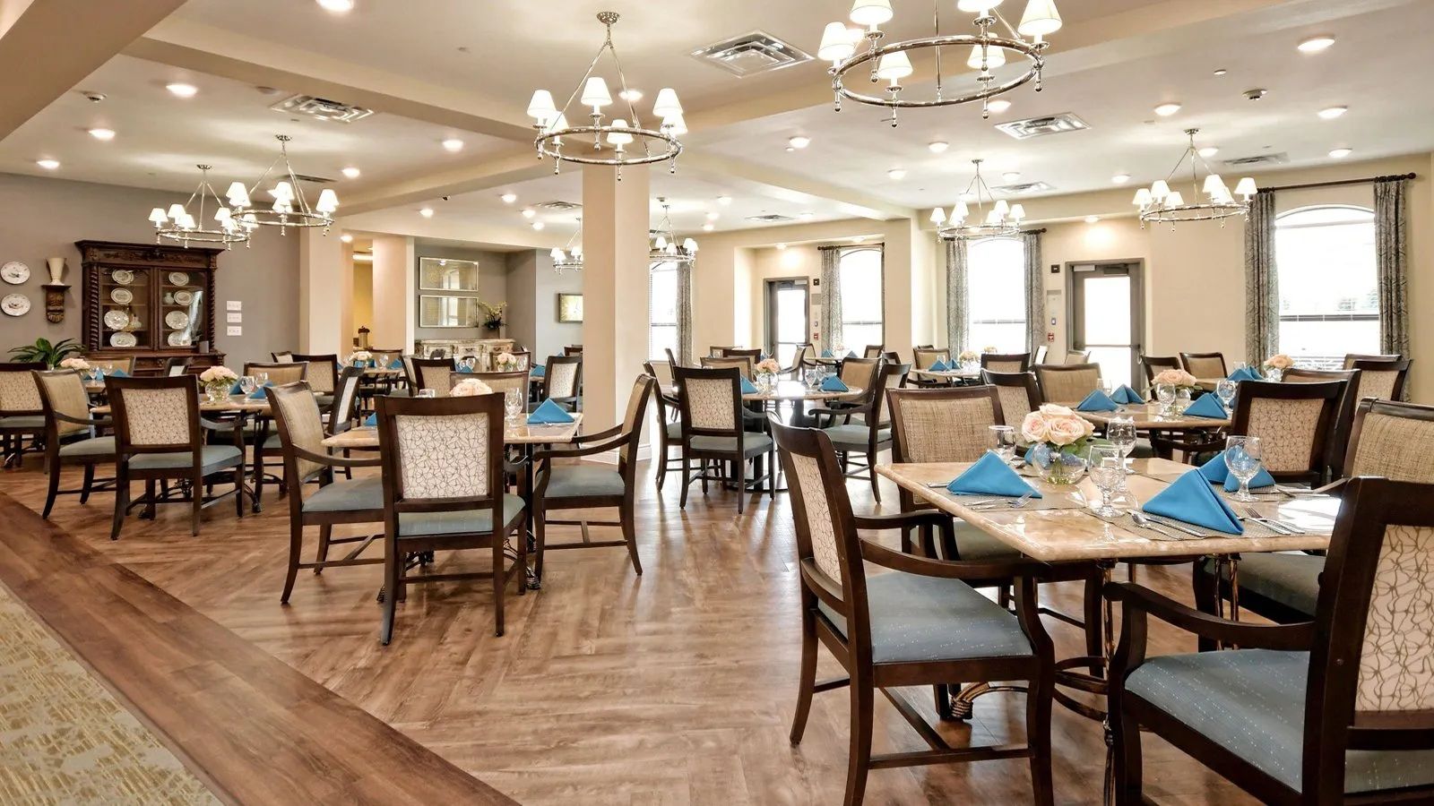 Elegant dining room with wooden chairs, round tables, chandeliers, and bright windows
